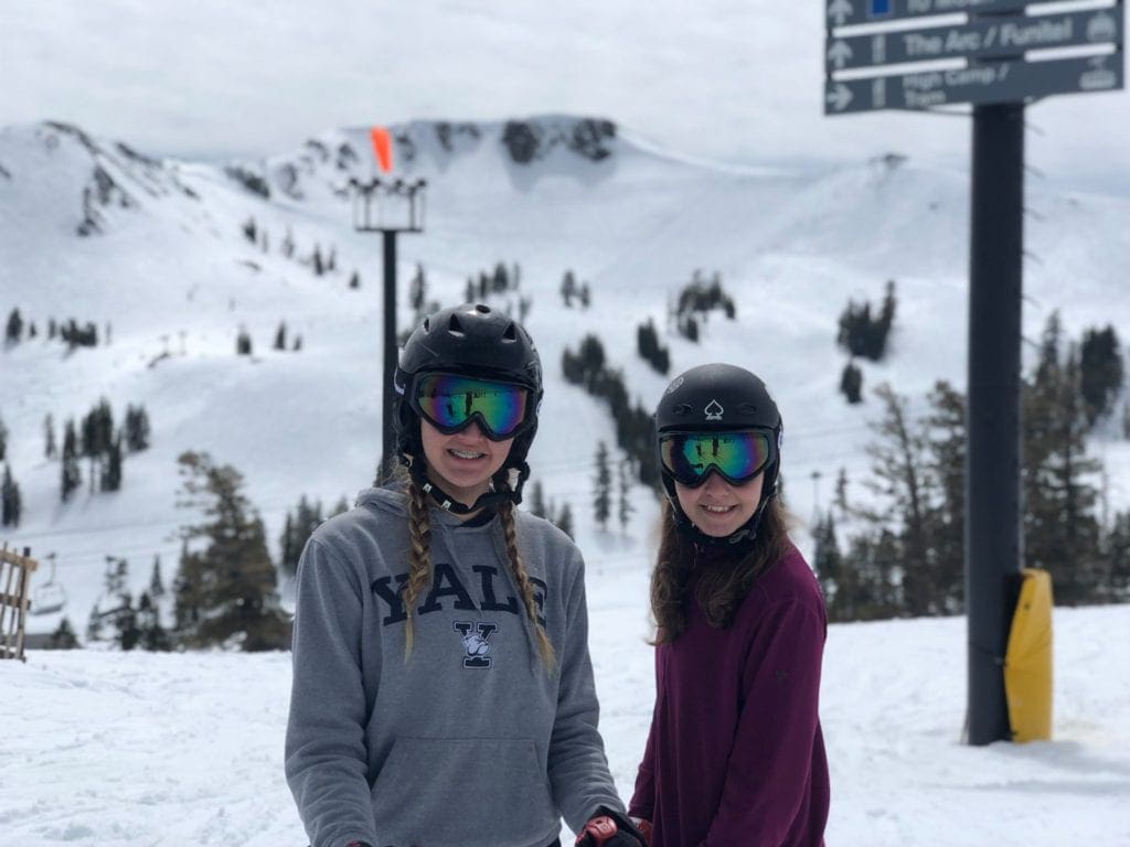 Two teen girls stand with sweatshirts, helmets, and snow goggles on with the slopes in the background at Squaw Valley, one of the best ski resorts near Lake Tahoe for families.