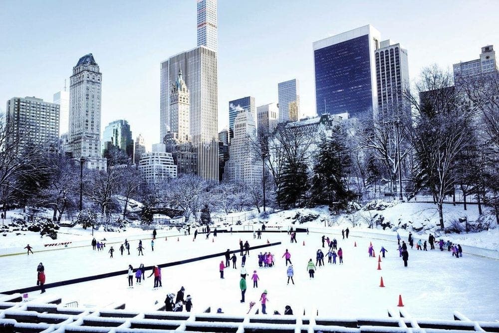 A view of the Wollman Rink on a snow-covered day featuring a number of skaters, and the NYC skyline behind them, one of the best New York City Christmas activities with kids.
