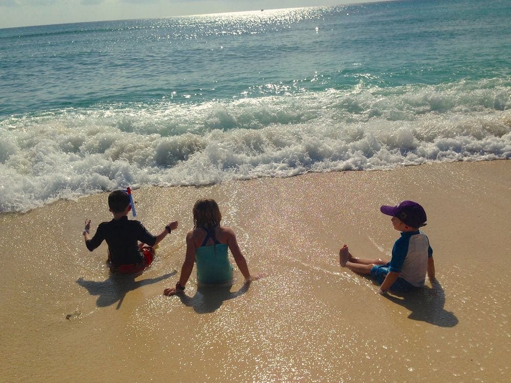 Three kids sit on the beautiful Cemetary Beach, one of the best things to do in Grand Cayman with kids.