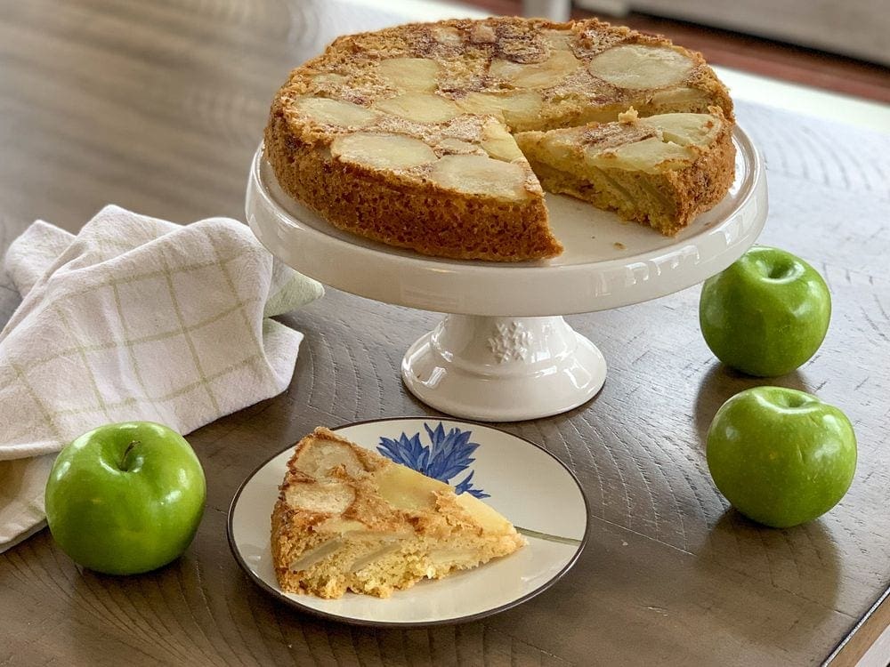 A slice of apple sharlotka sits near the rest of the cake on a cake stand surrounded by green apples.