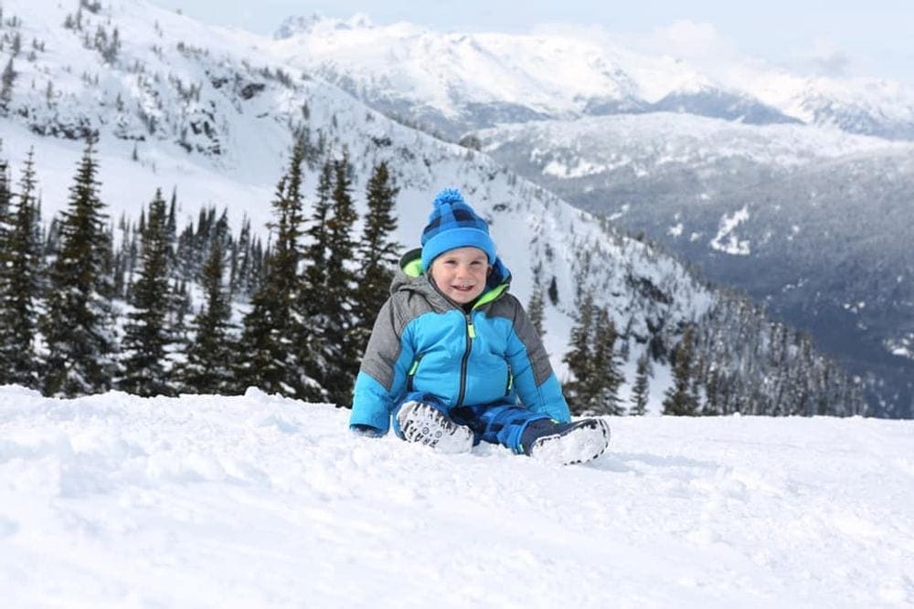 A young boy sits in the snow, wearing a blue coat and hat, in British Colombia.