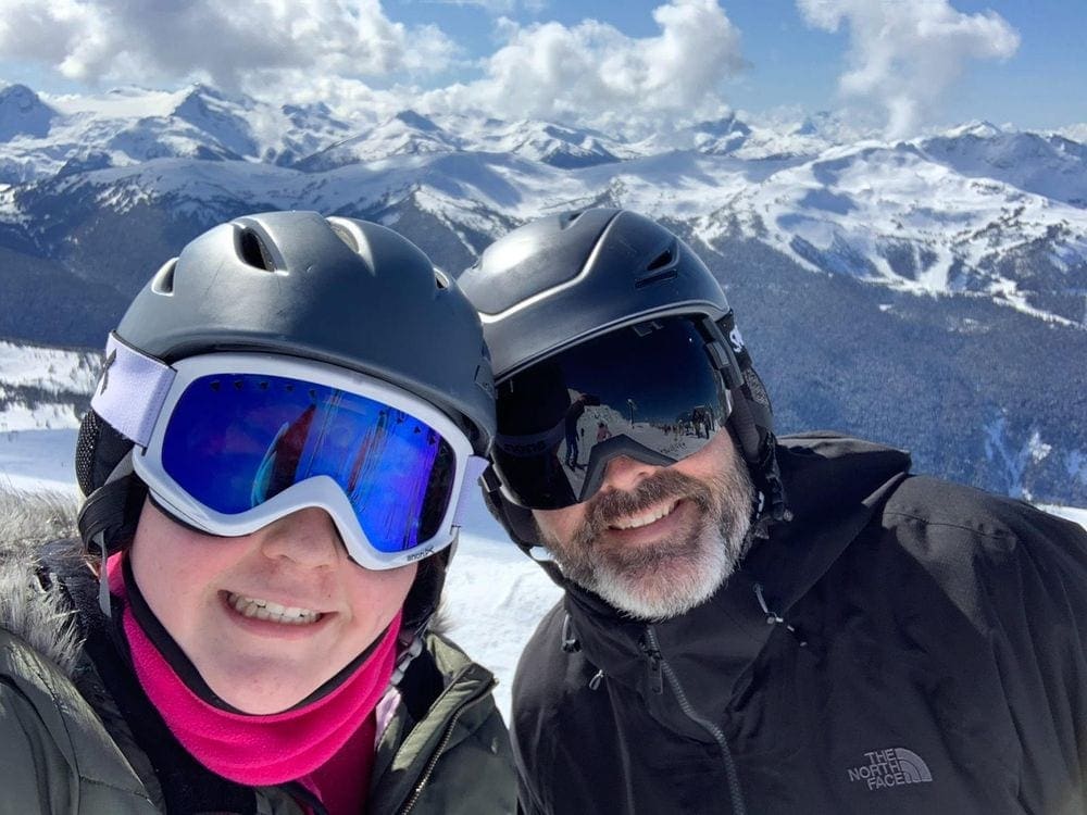 Two adults smile while wearing helmets and ski goggles on the slopes in Whistler.
