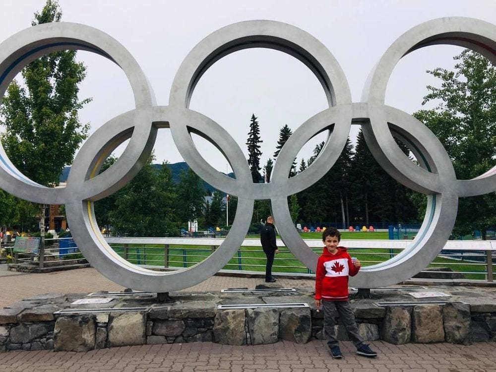 A young boy wearing a sweatshirt with a Canadian maple leaf on it stands in front of the Oylmpic statue at Whistler.