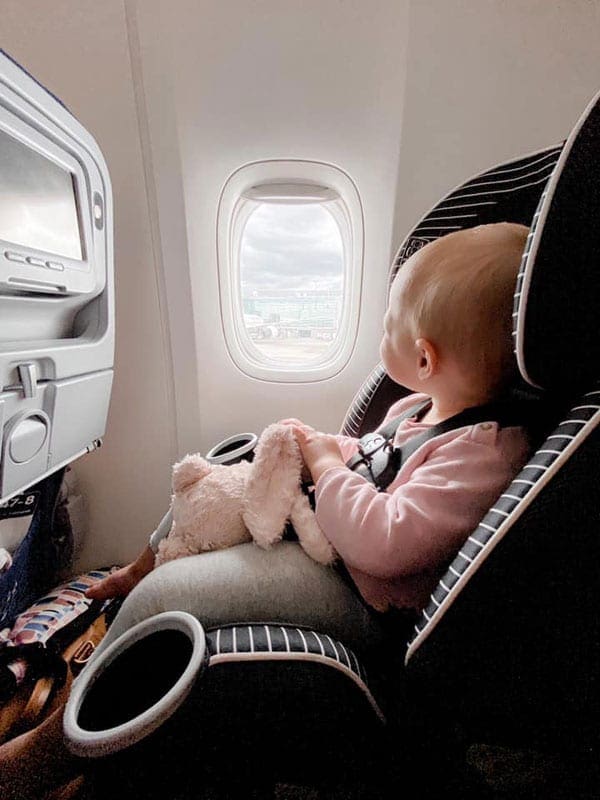 Infant in pink and grey outfit holding pink stuffed animal sitting in carseat on an airplane looking out the window