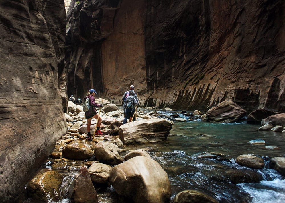 Three people walk among large boulders along a river in the Narrows, a must stop on your Utah road trip itinerary!