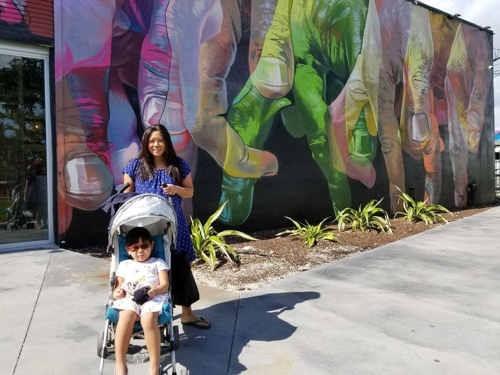 A mom stands behind a stroller, where her daughter sits, in front of a colorful art installment at Wynwood Walls, one of the best things to do in Miami with kids!