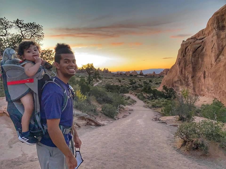 A man of color smiles while holding his young son in a backpack on a hike in Arches National Park, one of the best national parks for families.