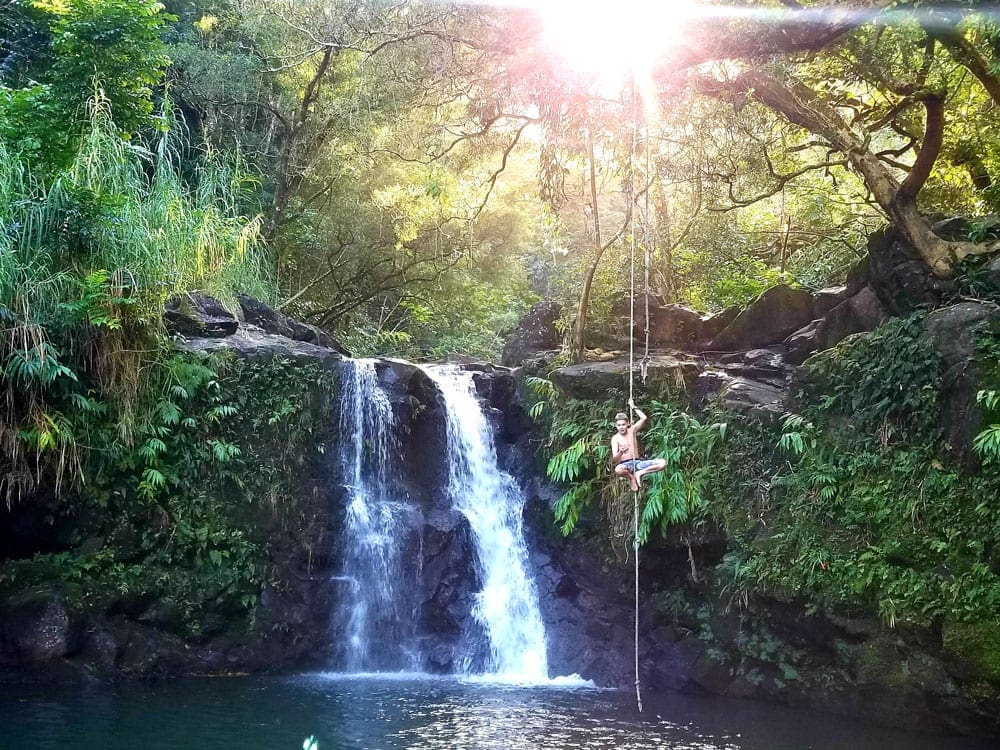A young boy climbs a roap over a Hawaiian pool and waterfall.