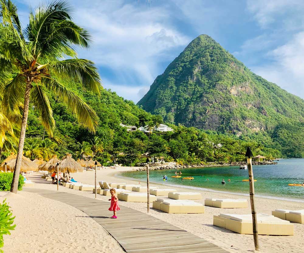 A toddler wearing red wanders along a boardwalk in St. Lucia, with the Pitons behinder her.