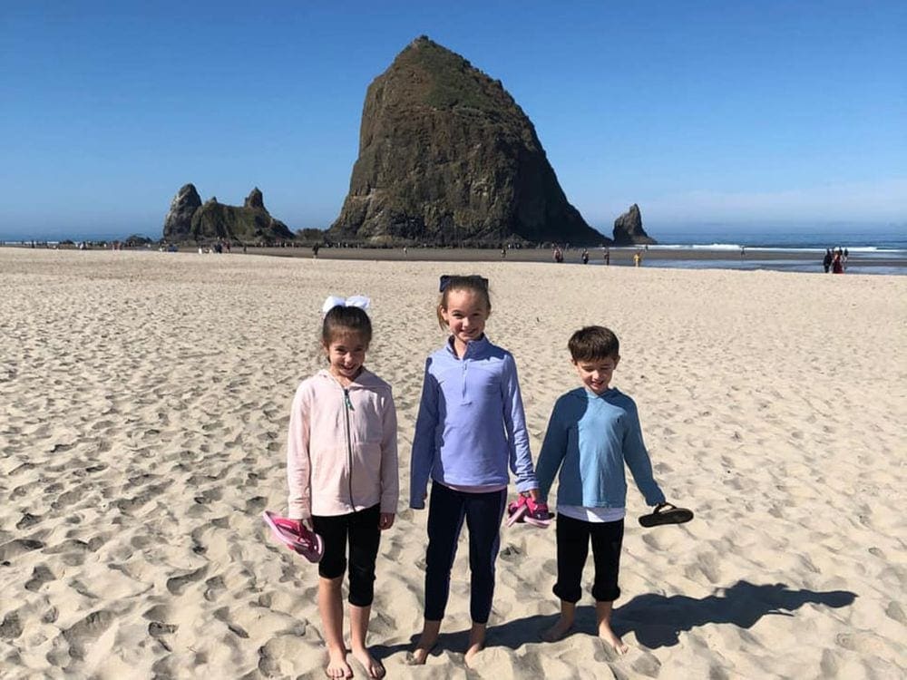 Three kids stand near one another on the beach with a large rock formation behind them at Cannon Beach in Oregon.