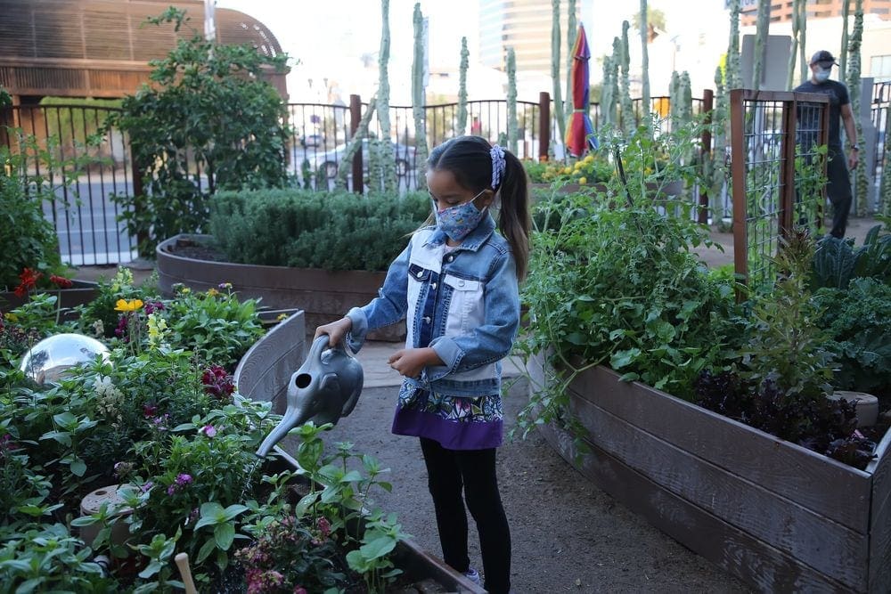 A young girl wearing a mask waters plants in the outside exhibit at the Children's Museum Phoenix, one of the best things to do Phoenix with kids.