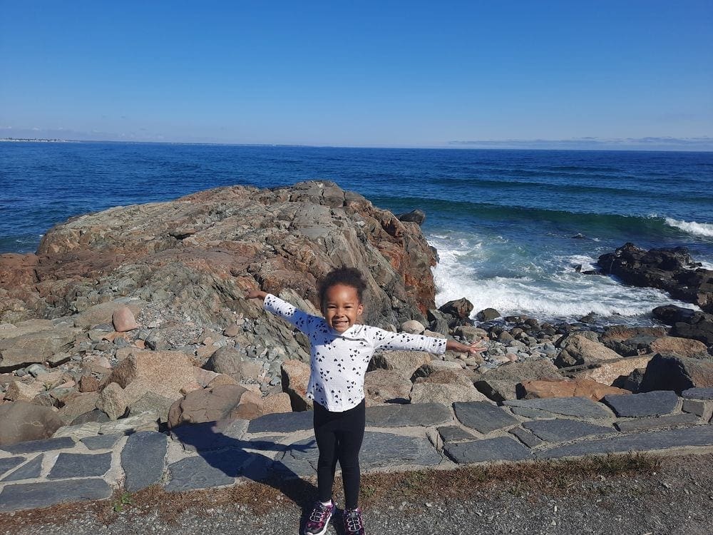 An African American toddler girls holds her arms wide open with a huge smile on her face, with Ogunquit Beach in Maine behind her, one of the best East Coast summer destinations for families.