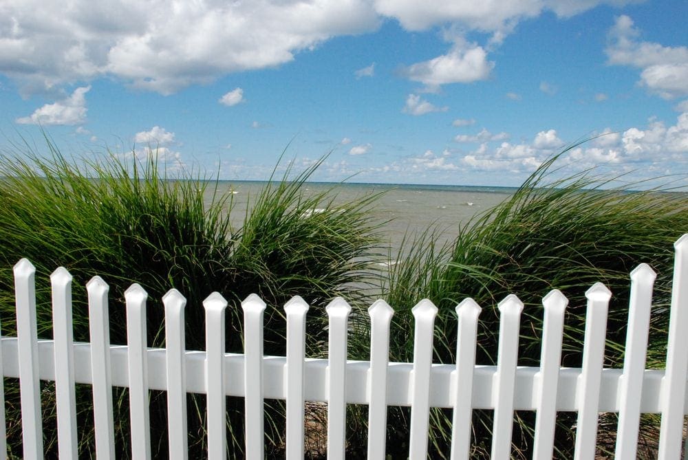 A white picket fence stands in front of tall grasses with Geneva On the Lake, one of the best lakes in the Midwest for families, in the background.