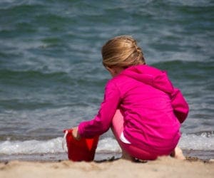 A young girl wearing a pink jacket plays with a red bucket on the beach.