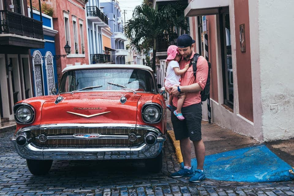 Dad holding a baby in old San Juan next two a red car.