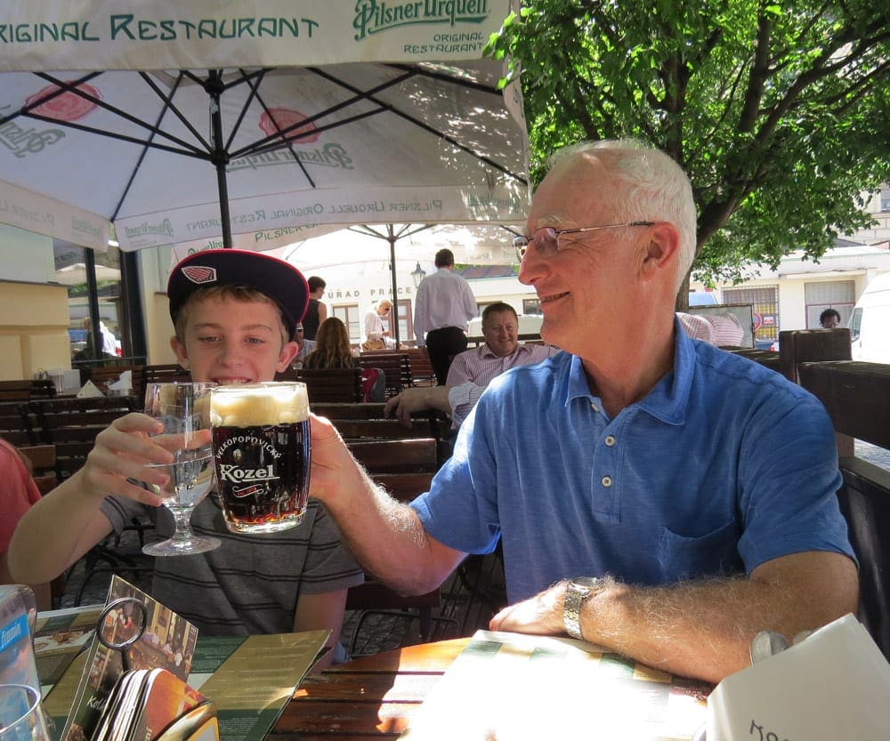 An older man holding a Czech beer and a young teen holding water cheers while exploring Prague on an Adventures by Disney tour.