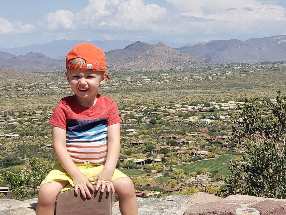 A young boy with a red cap on backwards sits on a stone fence with a desert view behind him while exploring Pinnacle Peak Park near Phoenix, one of the best places to visit in the US during Easter Break with kids!