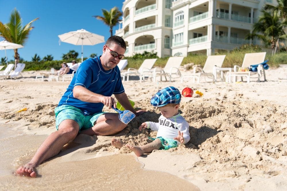 A dad and his infant son play int he sand on a sunny day at The Shore Club in Turks & Caicos, one of the best hot places to visit in December with kids!