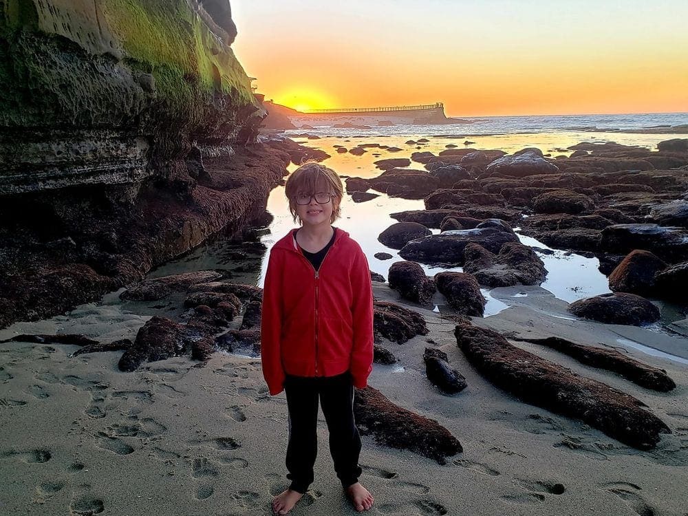 A young boy with glasses and a red shirt stands on the beach at La Jolla with large rock formations behind him at sunset.