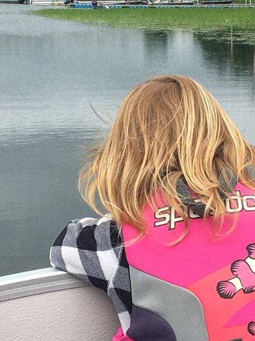 A young blond girl wearing a pink life jacket peers over the edge of a pontoon looking into Leech Lake.