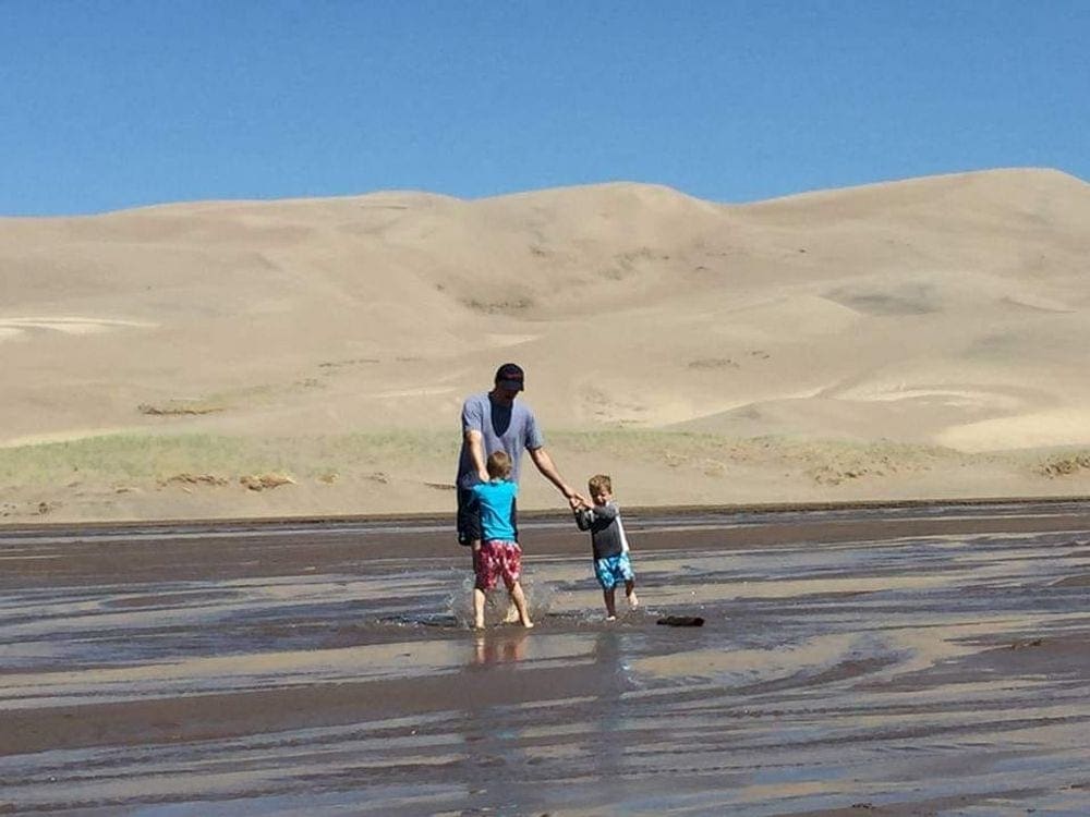 A dad dances with his two boys in the water at Medano Creek, one of our recommended kid-friendly U.S. beaches for families, with large sand dunes in the background.