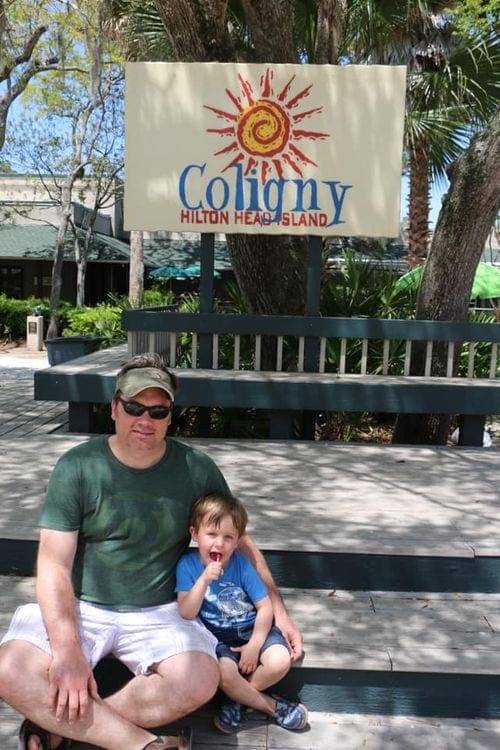 A father and young boy sits on a bench under a sign reading "Coligny, Hilton Head Island".