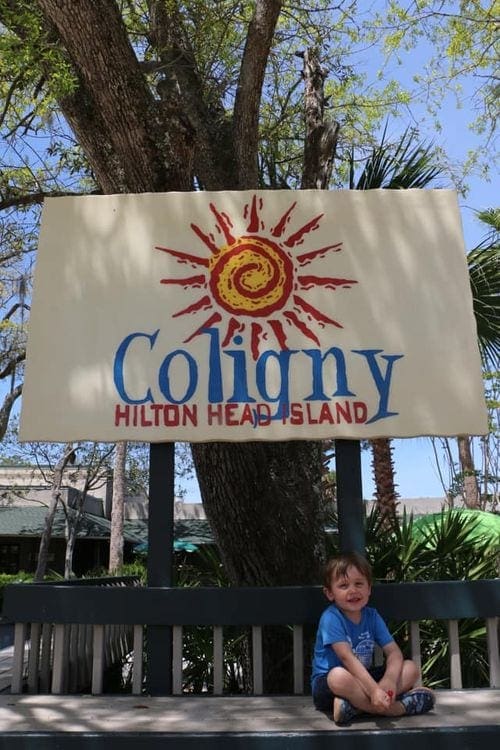 A young boy sits on a bench under a sign reading "Coligny, Hilton Head Island".