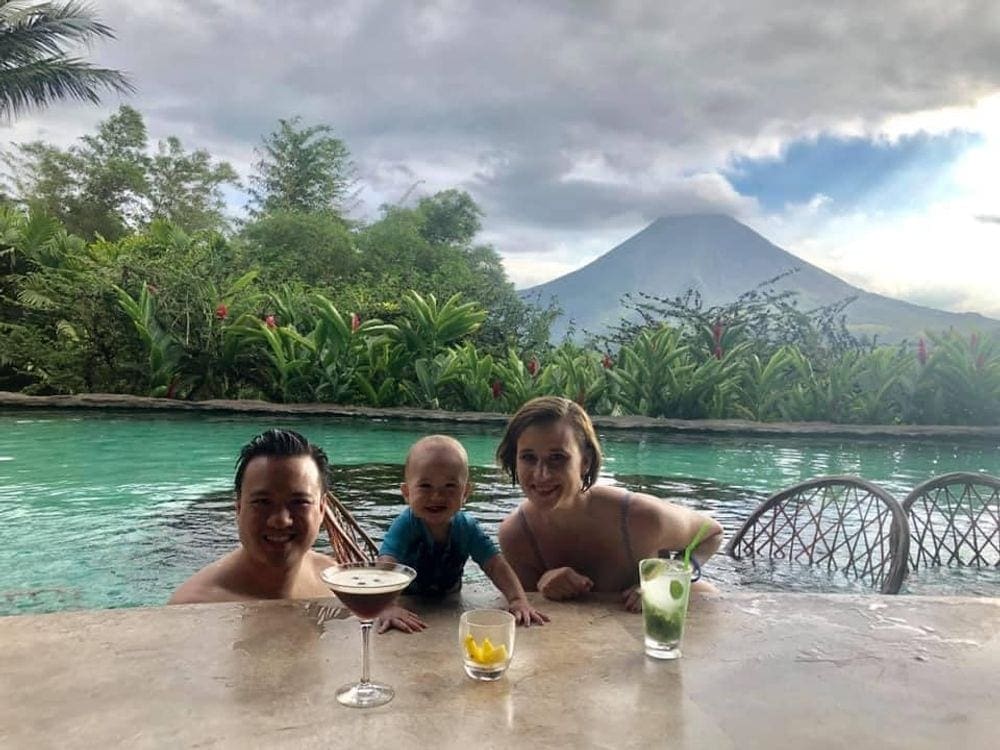 Two parents and a young baby enjoy time in a pool with the Arenal Volcano in the distance.