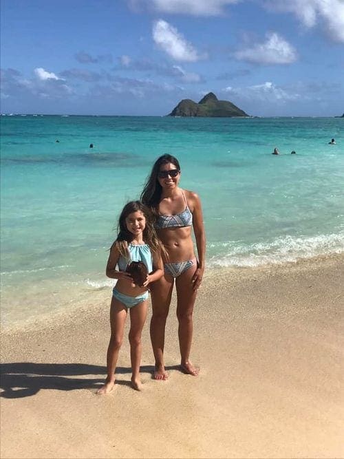 A mom and young daughter in bathing suits stand on Lanikai Beach on a sunny day.