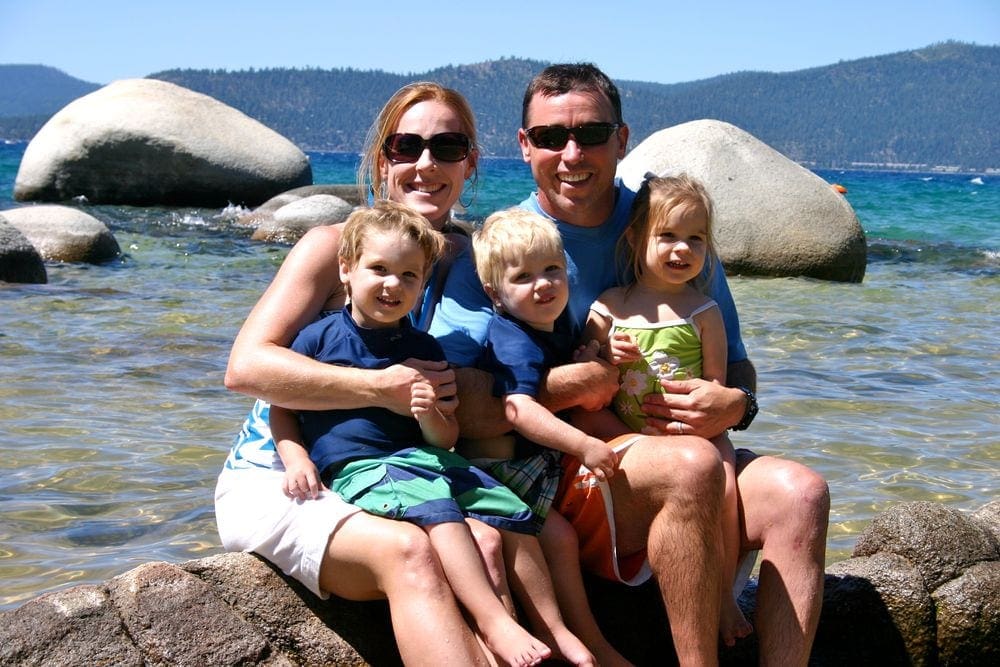 A mom and dad sit smiling with their three kids at Sand Harbor beach, one of our recommended kid-friendly U.S. beaches for families, large rocks nestle in the water behind them.