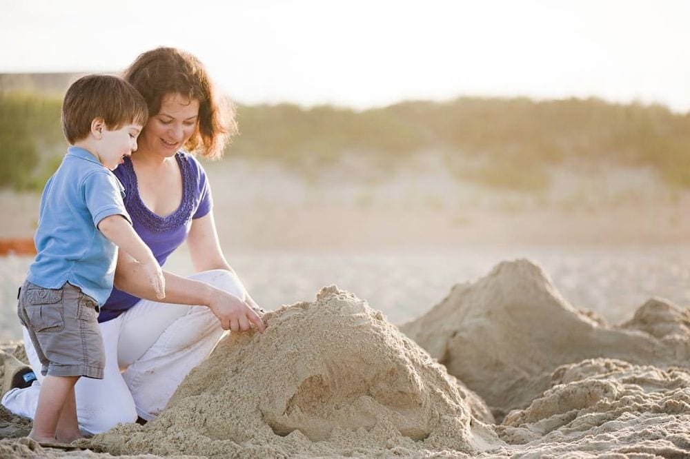 A mom and toddler son build a sand castle on a beach in the Outer Banks while staying at the Sanderling Resort Oasis Suites Hotel.