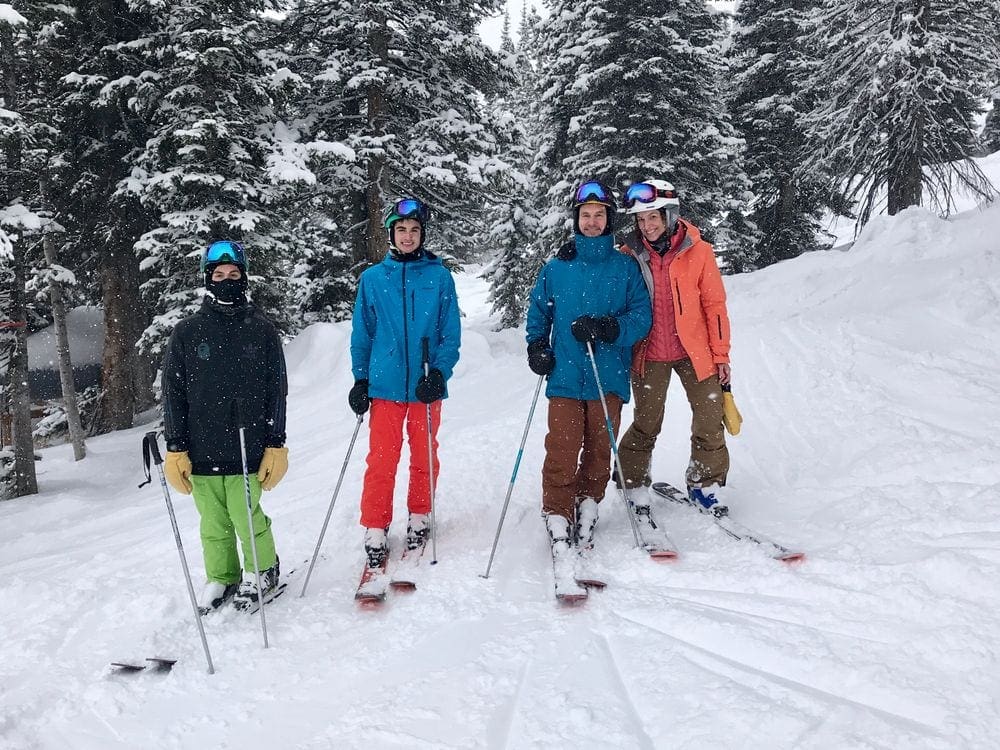 A family of four wearing skis and ski gear smiles at the camera while skiing at Jackson Hole, one of the best vacation spots in the US to impress teens and tweens.