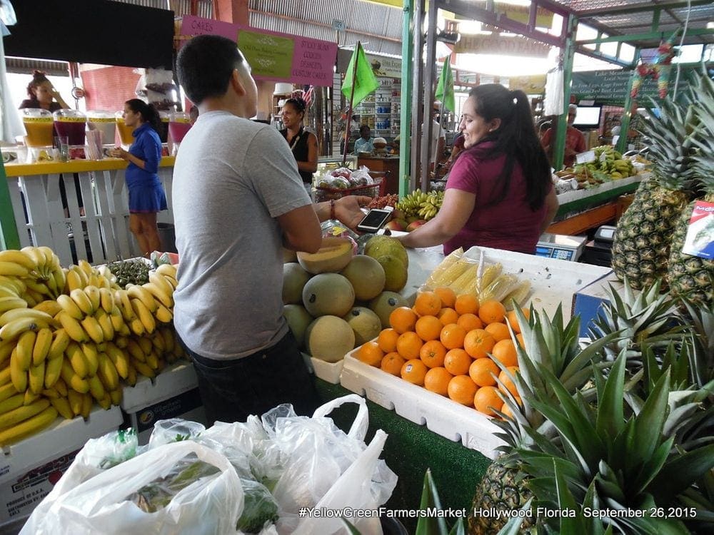 A man interacts with a vendor at the Yellow Green Farmers Market, while surrounded by fresh fruit.