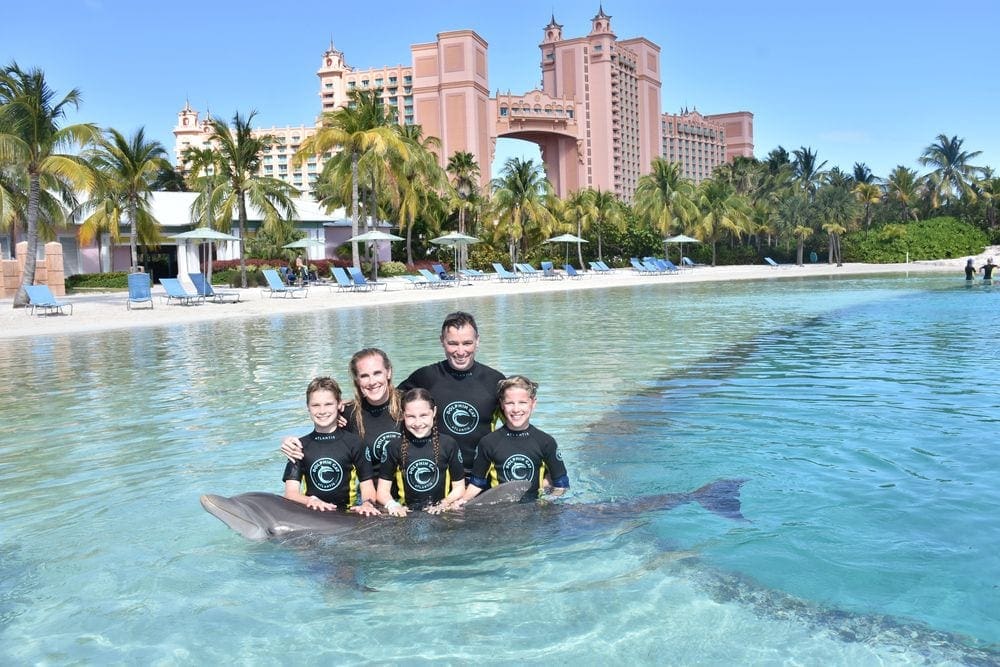 A family of five stands with a dolphin, with the resort buildings for Atlantis Bahamas in the background.