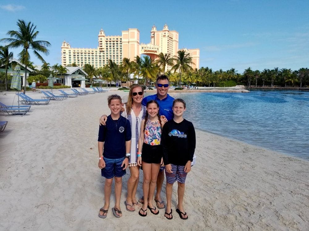 A family of five stands smiling on a beach in front of the iconic towers for the Atlantis Paradise Island, Bahamas, one of the best hotels in the Bahamas for families.