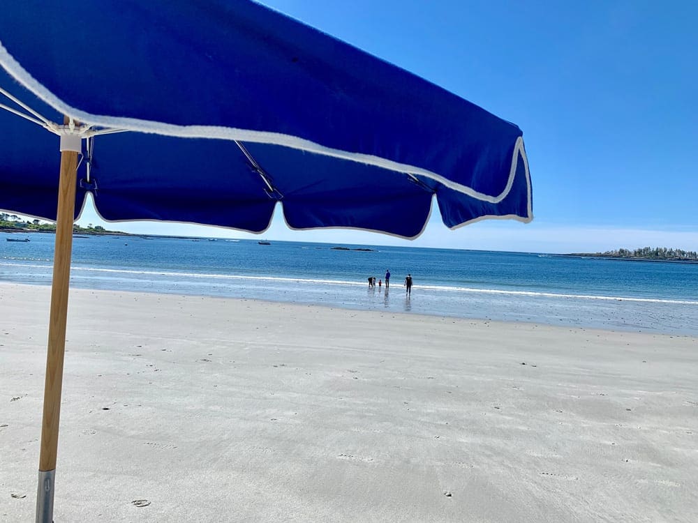 A beach umbrella shades an onlooker who sees a family wandering the beach near Inn by the Sea, one of the best eco-friendly hotels in the United States for families.