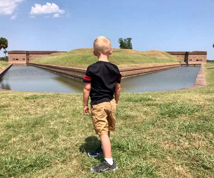 A young boy explores Old Fort Jackson on a sunny day while looking at the moat around the buildings.