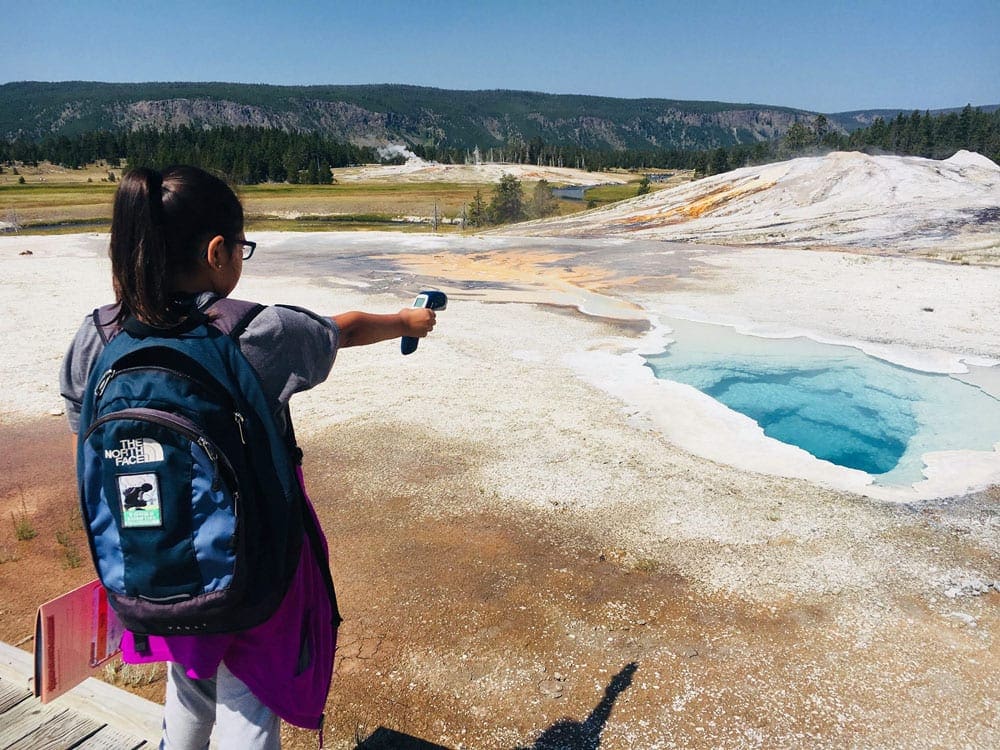 A young girl holds out a thermometer testing the temperature at a gyser in Yellowstone National Park, one of the best Summer Vacation Ideas in the U.S. for Families.