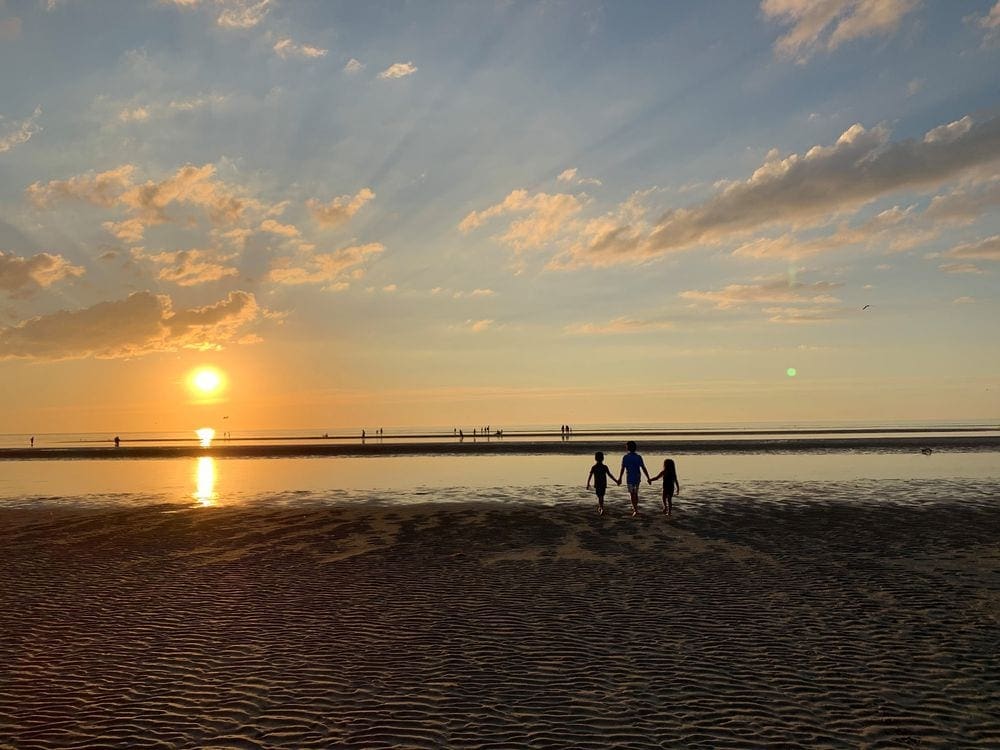 A mom holds her children's hand while walking on a beach in Cape Cod at sunset.