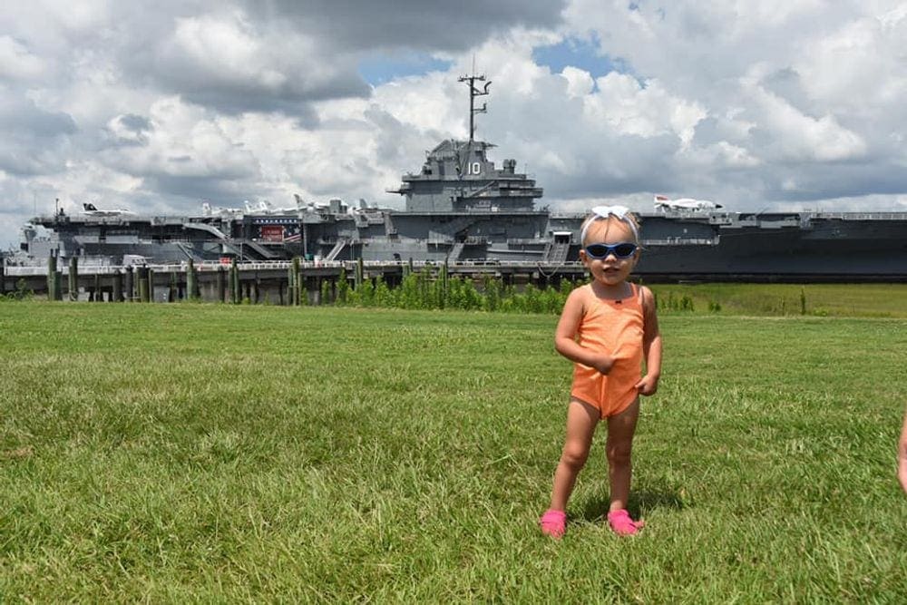 A young toddler wearing sunglasses stands in front of a large battleship.