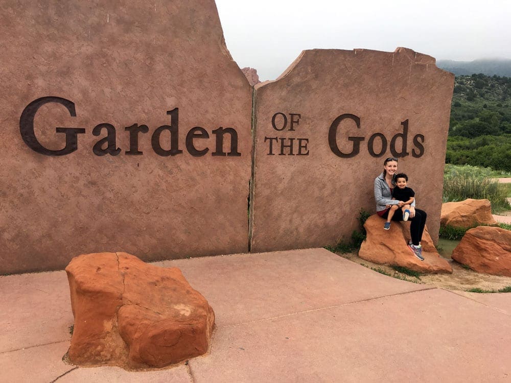 A mom holds her son while sitting on a rock in front of the sign for Garden of the Gods in Colorado, one of the best Summer Vacation Ideas in the U.S. for Families.