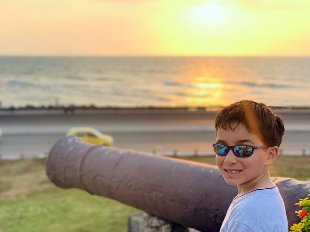 A young boy wearing sunglasses smiles while standing near a cannon on the Old Wall of Cartagena, with a stunning sunset over the water.