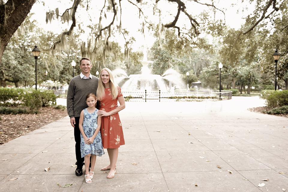 A family of three stands together smiling in front of the inconic fountain in Savannah.