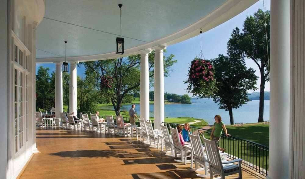 Several people, including kids, enjoy pristine white chairs on a balcony overlooking the ocean from the veranda of the The Otesaga Resort Hotel.