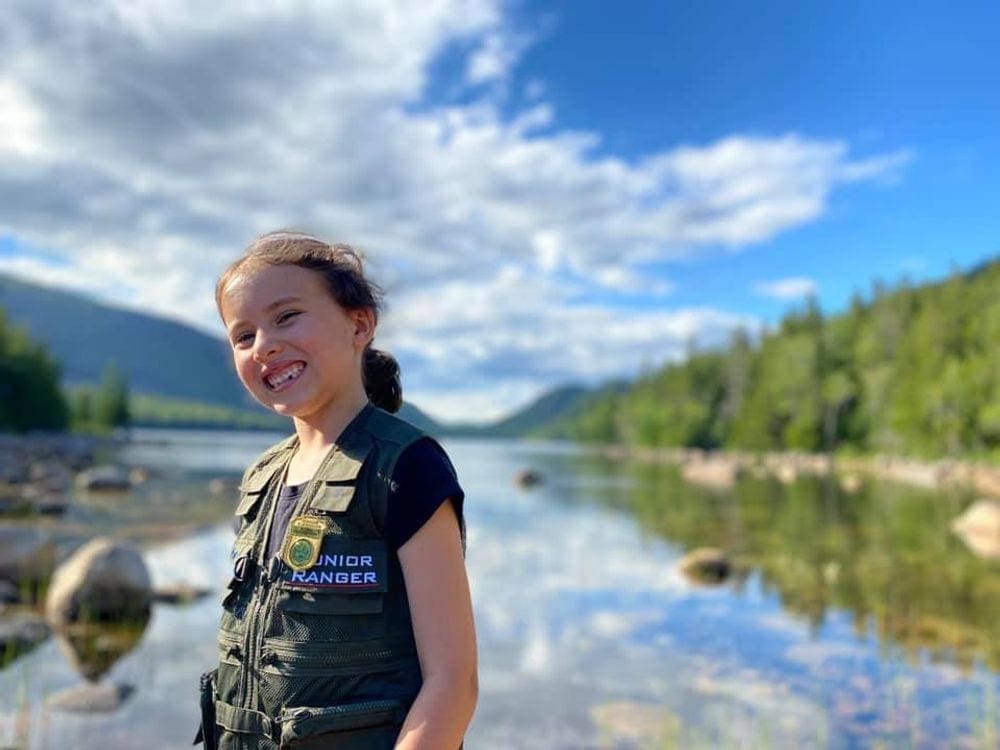 A young girl smiles broadly on a sunny day while explore Jordan's Pond in Acadia National Park, one of the best affordable summer vacations in the United States with kids.