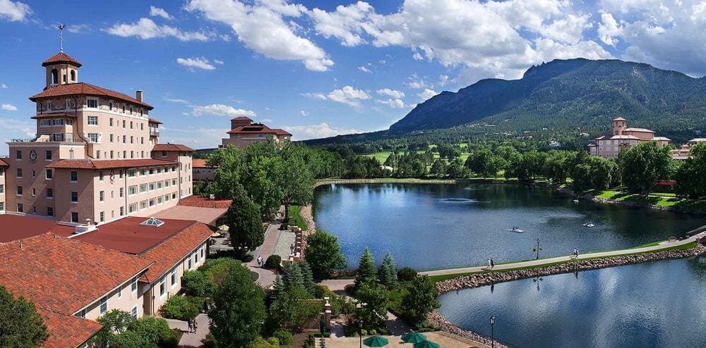 The Broadmoor Resort, large resort building on the left with a beautiful lake nestled on the right side and mountains in the distance.