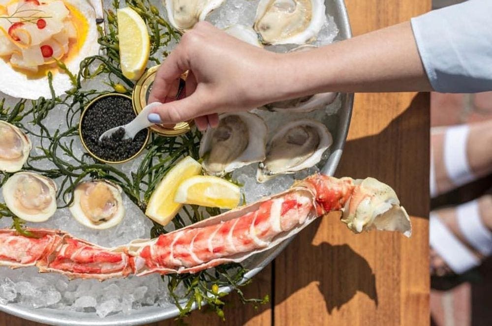 A hand reaches out to scoop cavier on a dish filled with lemon, oysters, and crab legs on an outdoor patio at Cru Nantucket.