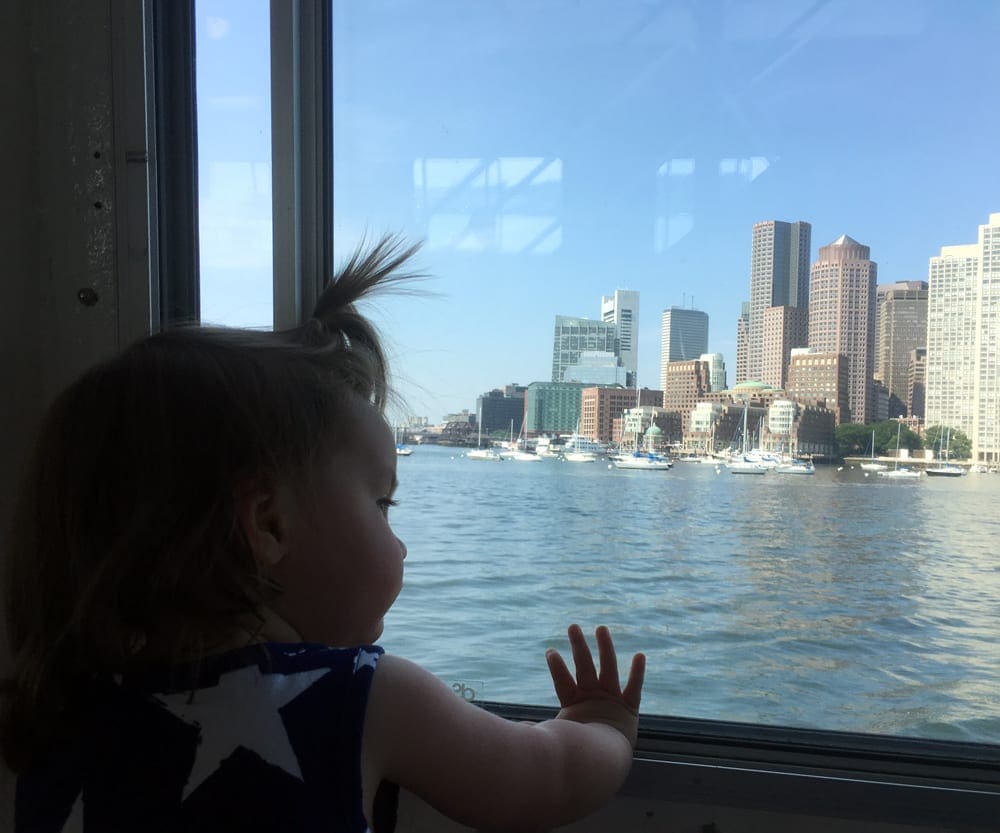 A baby looks out a window at the skyline of Boston from the water.