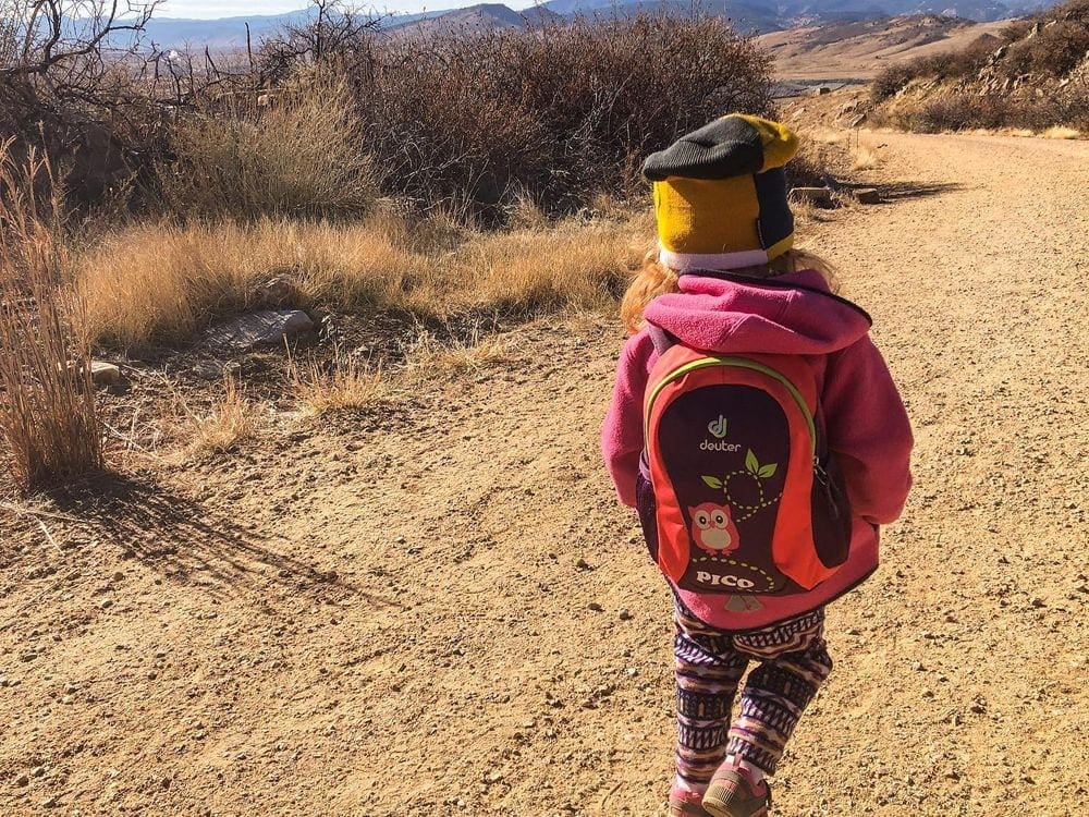 A young girl wearing a Deuter backpack, featuring an owl on it, walks along a hiking trail, one of the best gifts for travelers.