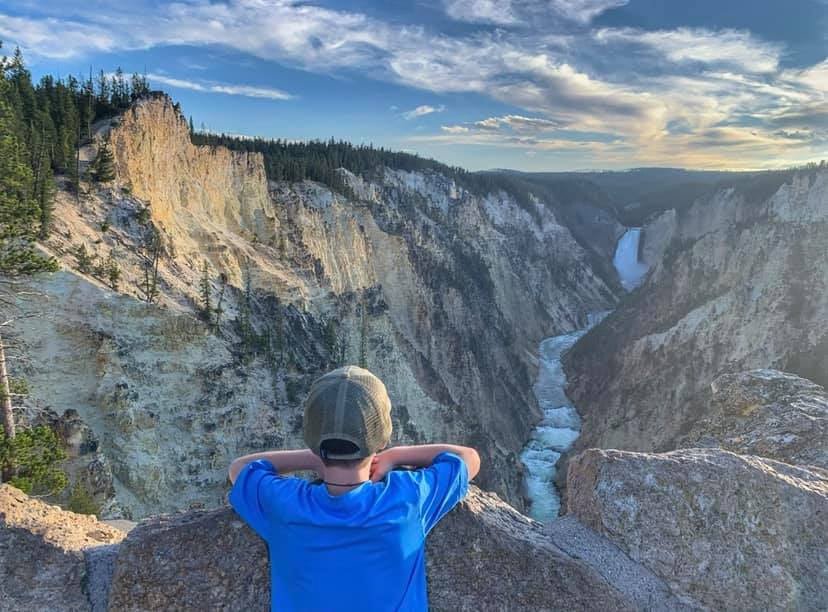 A young boy wearing a blue shirt looks over a scenic outlook of Yellowstone National Park, featuring a large waterfall.